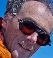 Cropped head portrait of Mr Stephen Venables a recent speaker. There is a blue sky behind him and he is wearing mountaineering sunglasses and an orange mountaineering sun glasses